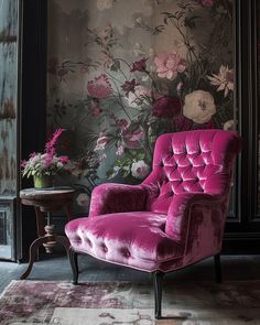 a pink chair sitting in front of a wall with flowers on it and a table next to it