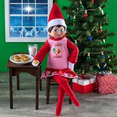 an elf sitting in front of a christmas tree with cookies on the table next to it