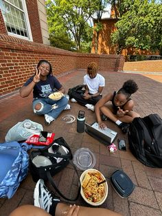 four people sitting on the ground eating food and talking on their cell phones with backpacks