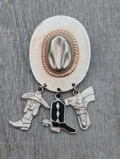 This fun brooch features a three dimensional silver tone cowboy hat that has a very slight warm, rosy wash. Three silver tone charms dangle from the hat's brim. One is a hound dog wearing its own hat and nestled in a cowboy boot. One is a boot with spurs and is covered in glossy black enamel. The third is a revolver in a decorated holster. This statement brooch is nicely weighted and wears very well. The piece is in great condition. It shows very minor wear consistent with vintage jewelry. The c Western Silver Hats For Western-themed Events, Western Hat Pins, Western Hat Pin, Silver Western Hat For Western-themed Events, Vintage Brown Jewelry For Rodeo, Vintage Silver Jewelry For Rodeo, Cowboy Hat Charm, Fun Brooch, Statement Brooch