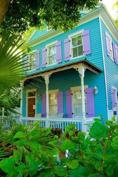 a blue house with purple shutters on the front porch and green trees around it