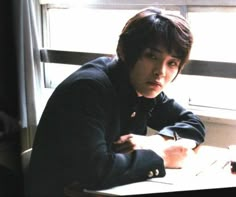 a young man sitting at a desk in front of a window with his arms crossed