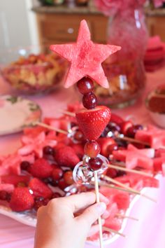 a person holding a stick with strawberries on it in front of watermelon and cherries