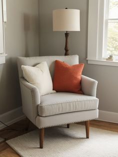 a white chair with an orange pillow on top of it in front of a window