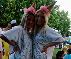 two women dressed up as unicorns pose for the camera at an outdoor event with other people in the background