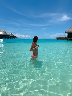 a woman standing in the ocean holding a cup and looking at the water with thatched huts in the background