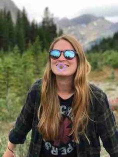 a woman with sunglasses on her face standing in front of some trees and bushes, blowing bubbles