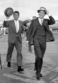 two men in suits and hats walking on an airport tarmac, one carrying a hat