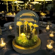 a table topped with a glass dome covered in yellow mushrooms and greenery next to lit candles