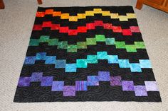 a black quilt with multicolored squares on it sitting on the floor next to a wooden table