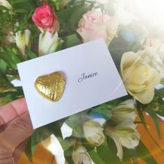 a person holding up a card with a gold heart on it and flowers in the background