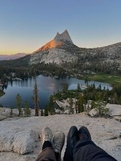 someone is sitting on the edge of a cliff overlooking a lake and mountains at sunset