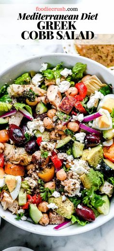 a white bowl filled with greek salad and topped with chicken, lettuce, tomatoes, cucumber, red onion, feta