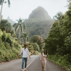 a man and woman walking down the middle of a road in front of some trees