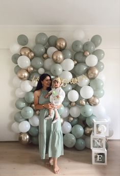 a woman holding a baby standing in front of a balloon wall with gold and white balloons