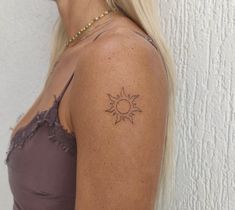 a woman with a small sun tattoo on her left shoulder and right arm, standing against a white wall