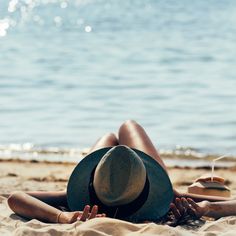 a woman laying on top of a sandy beach next to the ocean wearing a hat