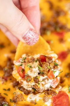 a hand holding a tortilla chip with meat and vegetables on it in front of a yellow casserole dish