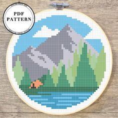 a cross stitch pattern with mountains and a lake in the foreground, on a wooden background