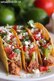 three tacos on a white plate with tomatoes, avocado and feta cheese