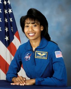 a woman in blue space suit sitting at a table with an american flag behind her