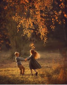 two young children holding hands under a tree in the fall time with leaves on it