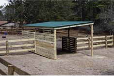 a horse pen with a green roof and wooden fenced in area next to trees