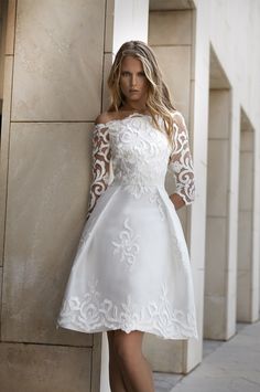 a woman is leaning against a wall wearing a white dress with lace detailing on the sleeves