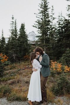 a man and woman standing next to each other in front of pine trees on the side of a road