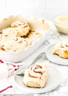 two plates with cinnamon rolls on them next to a baking pan and bowl of cream cheese