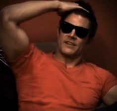 a man in an orange shirt and sunglasses sitting on a couch with his hands behind his head