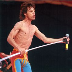 a man with no shirt on holding a microphone in his right hand and wearing colorful pants