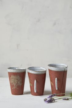 three cups sitting next to each other on top of a white table with purple flowers