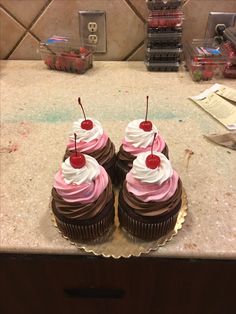 three cupcakes with pink frosting and cherries on top sitting on a counter