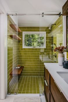 a green tiled bathroom with wooden cabinets and white counter tops, along with a window above the sink