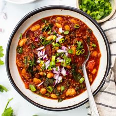 a white bowl filled with chili and beans next to some cilantro, green onions, and limes