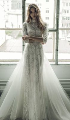 a woman standing in front of a window wearing a white wedding dress with long sleeves