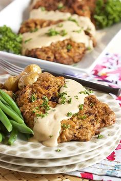 two white plates topped with meat covered in gravy next to green beans and broccoli