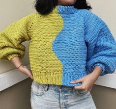 a woman wearing a yellow and blue sweater with her hands on her hips, standing in front of a window