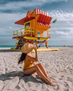 a woman sitting on the beach in front of a lifeguards house with a straw hat