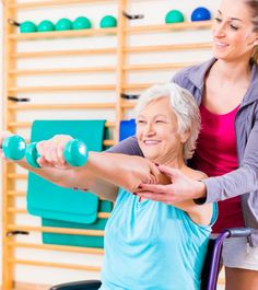 an older woman in a wheel chair holding two dumbbells while another woman stands next to her