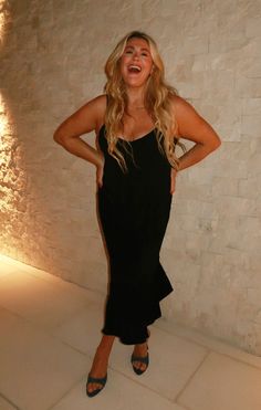 a woman standing in front of a white brick wall wearing a black dress and smiling