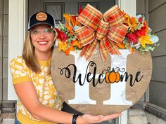 a woman holding up a welcome sign in front of a door with fall decorations on it