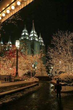 people are walking through the snow in front of lighted trees and buildings with lights on them