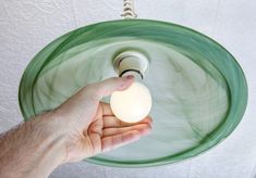 a hand holding a light bulb in front of a green glass bowl fixture on a white wall