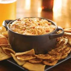 a bowl of dip surrounded by crackers and chips on a plate with beer in the background