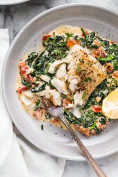 a white plate topped with fish and spinach covered in sauce next to a lemon wedge