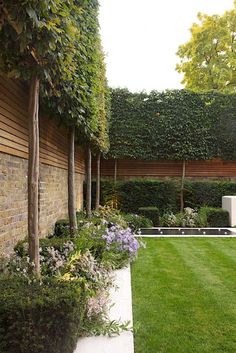 an outdoor garden with grass, shrubs and flowers on the side of a brick wall