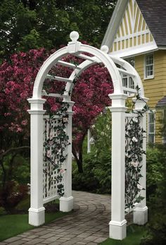 a white garden arbor with pink flowers on the top and green grass around it, in front of a yellow house