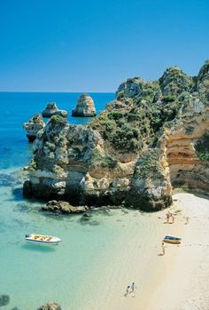 people are walking on the beach next to some rocks and boats in the clear blue water